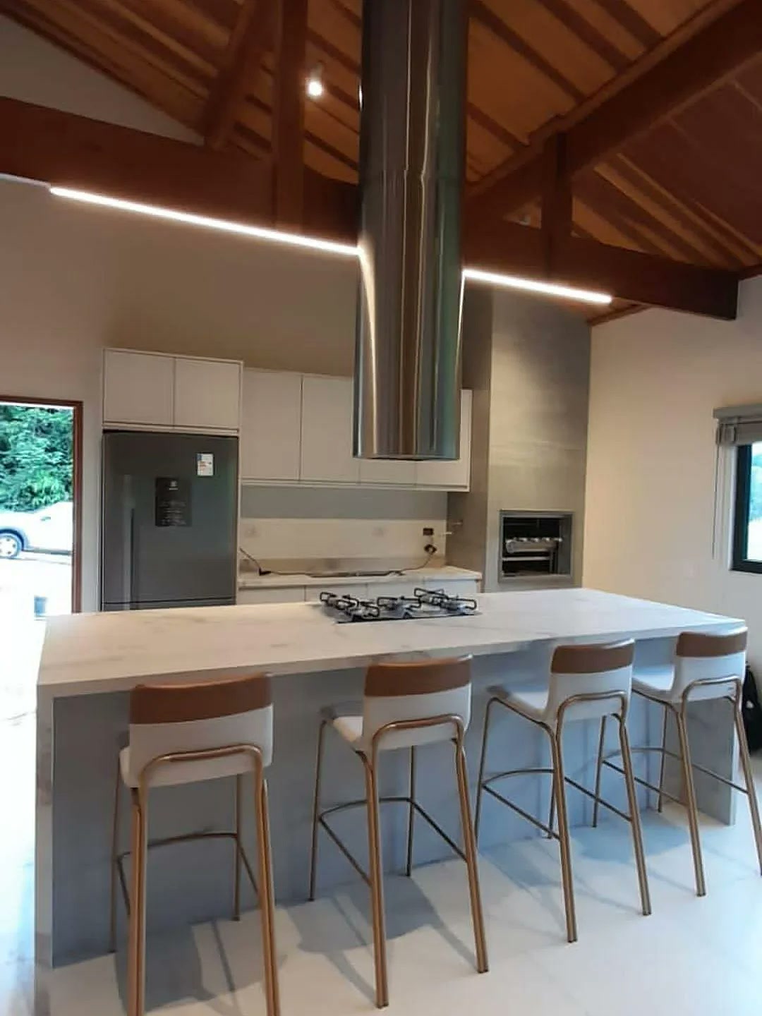 Cozinha gourmet planejada com ilha de mármore, coifa tubular, banquetas e teto em madeira – projeto Estillo Design